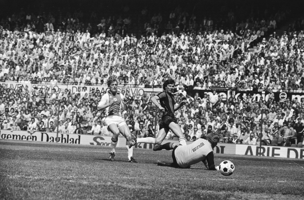 Schwarz-weiß-Foto von Männern, die auf einem Feld Fußball spielen, mit Zuschauern im Hintergrund und einer 'Bundesliga 1978-79 Ajax 1-1 Hannover 96'-Fahne.