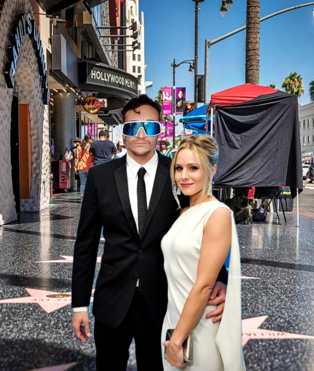 Ein Mann in einem schwarzen Blazer, weißem Hemd, schwarzer Krawatte und schwarzen Hosen steht neben einer Frau, die eine Geldbörse hält, auf dem Hollywood Walk of Fame, mit Menschen, Gebäuden, Bäumen, Pfählen, Laternen und Schildern im Hintergrund.