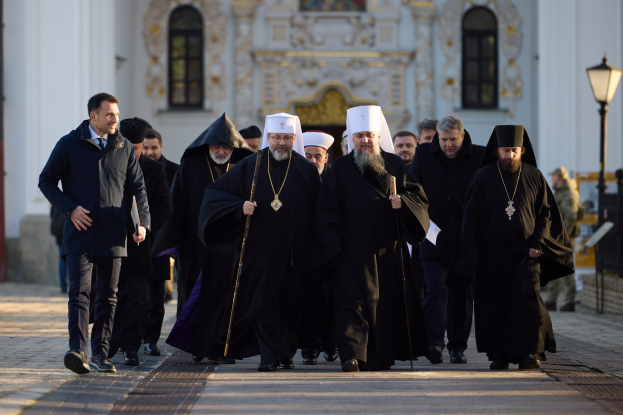 Eine Gruppe von Priestern, die über eine Straße gehen, an einem Gebäude mit Fenstern und einer Tür vorbei, einige tragen Käpchen und halten Stäbe, mit einem Laternenpfahl auf der rechten Seite und einigen Menschen im Hintergrund.