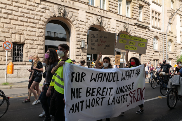 Eine Gruppe von Menschen marschiert mit Protestschildern und fährt Fahrräder, im Hintergrund ein klassisches Gebäude mit Rundbogenfenstern, Säulen und Reliefs.
