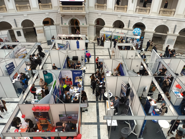 Große Gruppe von Menschen auf einer internationalen Fachmesse mit Ständen, Bannern, Tischen, Stühlen, Luftballons und einem Gebäude mit Bögen, Säulen, Geländern und einem Bildschirm im Hintergrund.
