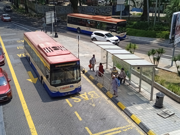 Stadtstraße mit fahrenden Autos, Bussen, einer Bushaltestelle, Fußgängern auf dem Gehweg, Strommasten, Schildern, Bäumen, Grünflächen und umliegenden Gebäuden.