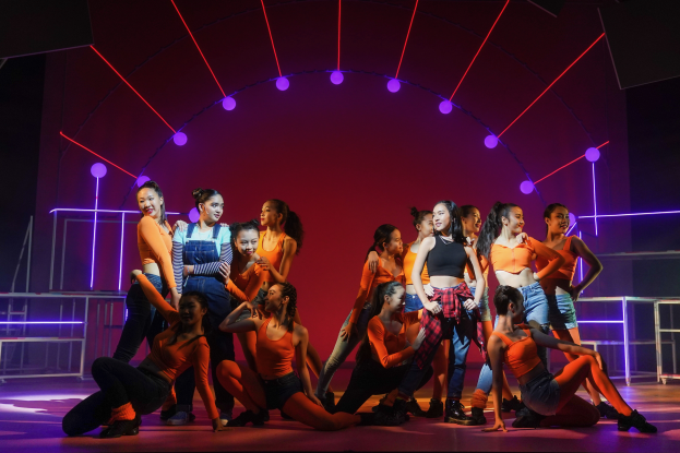 Eine Gruppe junger Tänzer in bunten Kostümen performs auf einer Bühne mit Geländern, beleuchtet von bunten Lichtern, mit einer jubelnden Menge im Hintergrund.