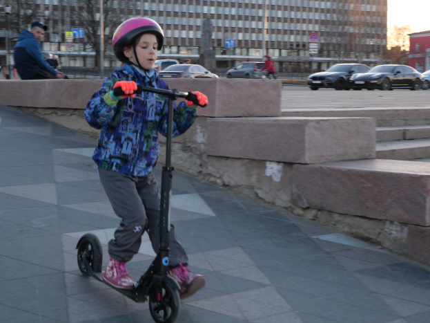 Ein junger Junge fährt mit einem Helm und Handschuhen auf einem Scooter einen Gehweg entlang, mit verschiedenen städtischen Elementen und einem klaren blauen Himmel im Hintergrund.