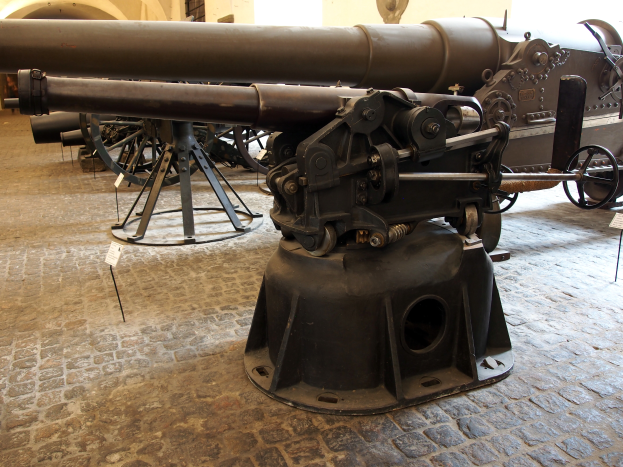 Eine große Gatling-Kanone mit langem Lauf und Seitengriff ist in einem Museum ausgestellt und von Bodenständen umgeben. Im Hintergrund ist eine Fensterwand zu sehen, die an eine Burg erinnert.