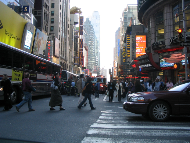 Eine belebte Stadtstraße mit Fahrzeugen, Fußgängern an einer Zebrastreifenkreuzung, Gebäuden, Schildern, Verkehrsampeln und einem klaren blauen Himmel.