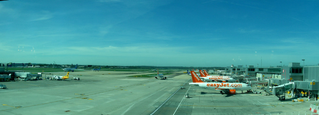 Eine Luftansicht eines Flughafens mit Flugzeugen, Gebäuden, Polen, Fahrzeugen, Bäumen, einer Straße unten und einem Zaun im Vordergrund.