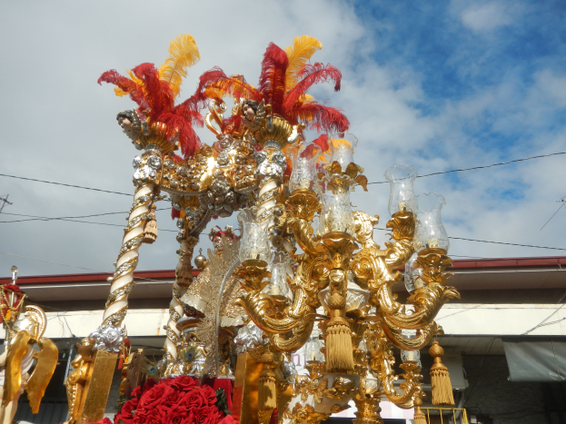 Ein großes goldenes und rotes Festwagen, das mit Blumen und anderen Dekorationsgegenständen geschmückt ist, bei einem Karnevalsumzug, mit einem Gebäude, Strommasten mit Drähten und einem bewölkten Himmel im Hintergrund.