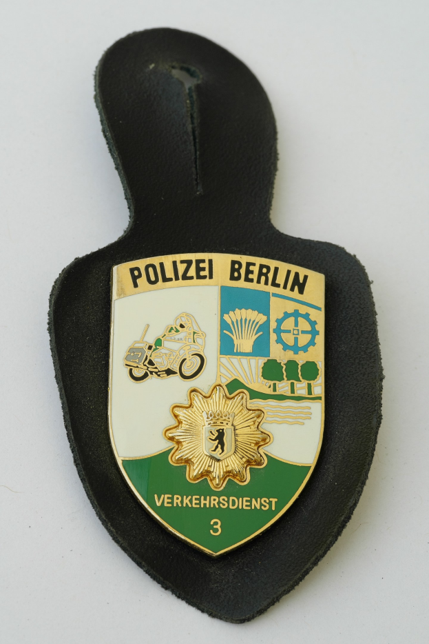 Ein schildförmiges Polizeibadgebilde mit "Polizei Berlin" in schwarzer Schrift auf einem blauen Hintergrund mit einem weißen Stern und Rand, das auf einer Oberfläche ruht.