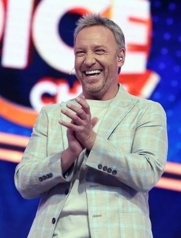 Ein Mann in einem Anzug klatscht auf einer Bühne mit 'The Voice'-Schriftzug im Hintergrund.