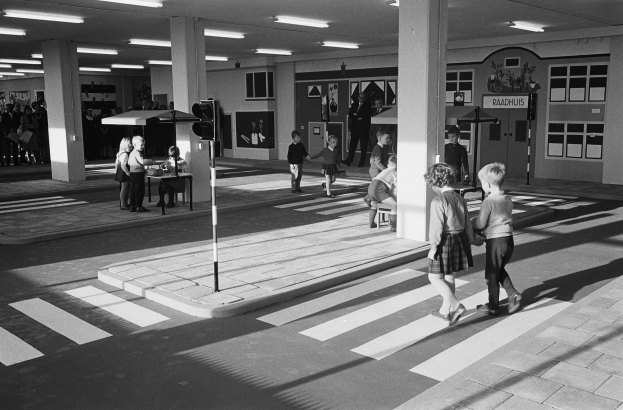 Ein Schwarz-Weiß-Foto von Kindern, die eine Straße an einer Ampel überqueren, mit Menschen, die auf Bänken stehen und sitzen, einem Verkehrssignal im Vordergrund und Säulen und Lampen im Hintergrund.