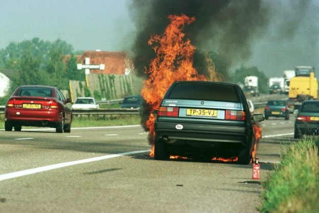 Ein Auto in Flammen an der Straße, mit anderen Fahrzeugen in der Nähe, Bäumen, Gebäuden und einem klaren blauen Himmel im Hintergrund und einem Feuerlöscher auf dem Gras rechts.