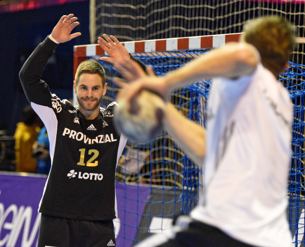Zwei Männer spielen Handball vor einem Tor mit Netz und einem Banner mit Text im Hintergrund und einigen Menschen in einem Stadion.
