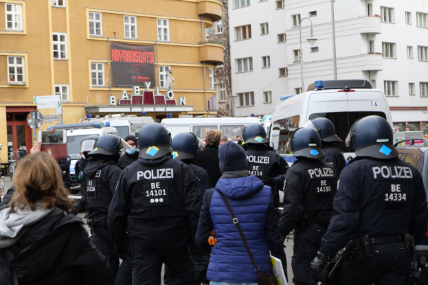 Polizeibeamte in Uniform vor einer Menge bei einer Demonstration in Berlin, mit Fahrzeugen, Gebäuden und einer Person mit einer Kamera im Hintergrund.