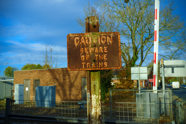 Warnschild an einem Bahnübergang mit Bäumen, Strommasten, einem Gebäude, Containern, Fahrzeugen und einer bewölkten Himmel.