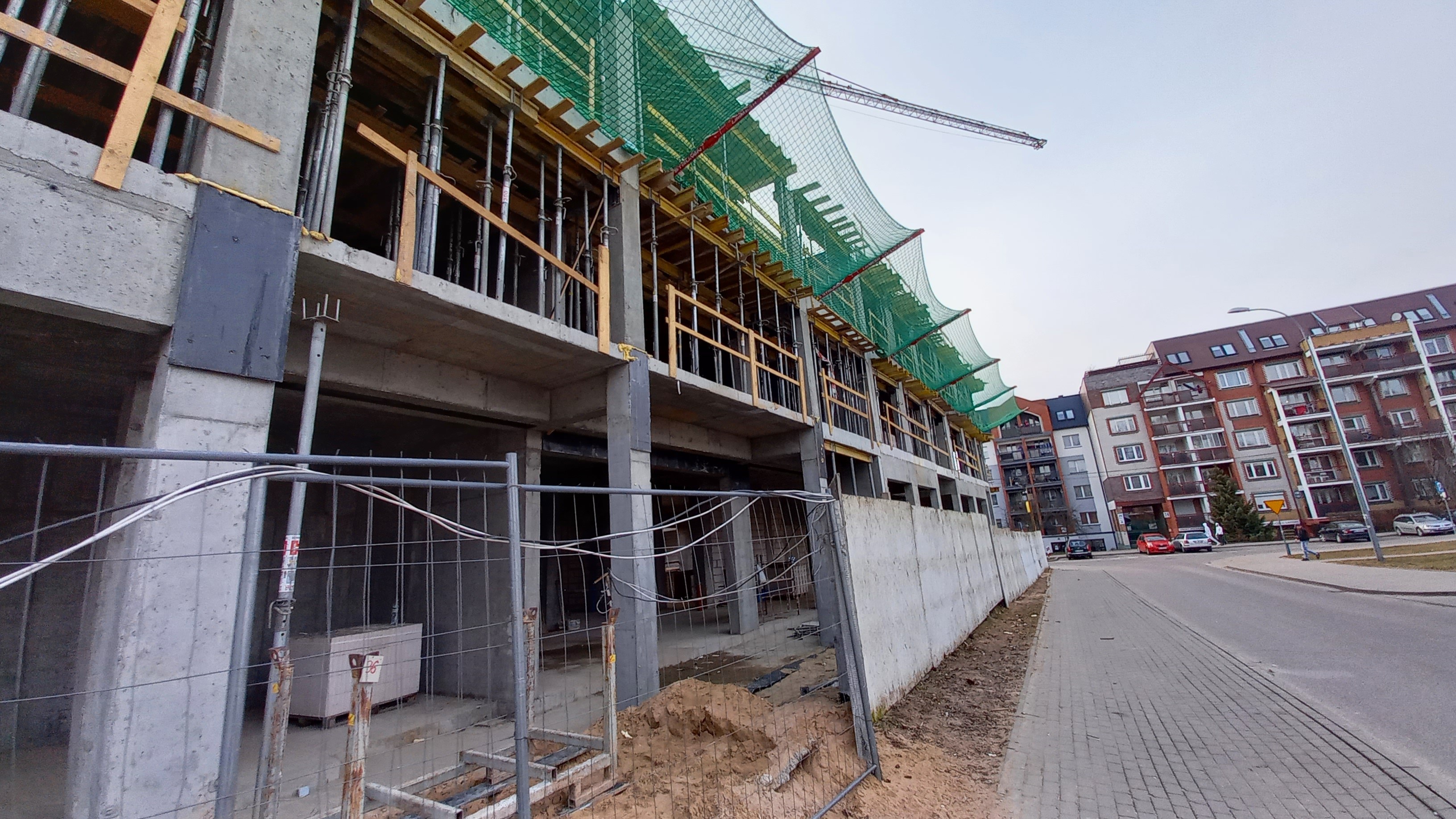 Großer Bau am Straßenrand, umgeben von Gebäuden, Straßenmasten, Straßenlaternen und Kraftfahrzeugen bei klarem blauem Himmel.