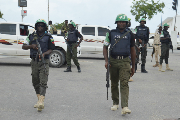 Eine Gruppe von Polizisten in Helmen und mit Waffen in der Hand geht eine Straße entlang mit Fahrzeugen, Laternenmasten, Verkehrsampeln, Schildern mit Text, Bäumen, Gebäuden und einem bewölkten Himmel im Hintergrund.