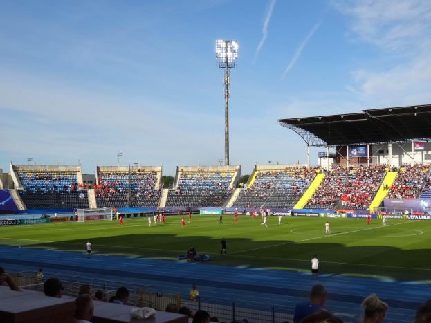 Ein Fußballspiel in einem großen, beleuchteten Stadion mit Zuschauern auf den Tribünen und Spielern auf dem Feld unter einem sichtbaren Himmel.