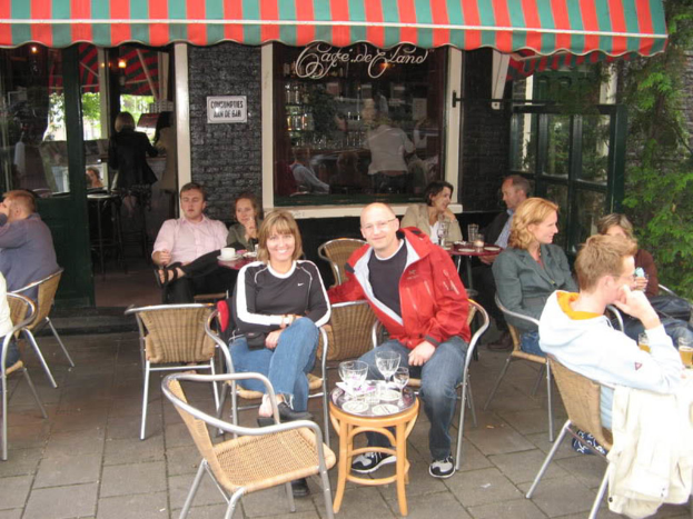 Eine Gruppe von Menschen sitzt um einen Tisch mit Tellern und Gläsern, lachender Mann und Frau in der Mitte, schwarze Backsteinwand, Fenster mit Text, Bäume, grüne Tür mit einer Frau drinnen und ein grünes und rotes Zelt darüber.