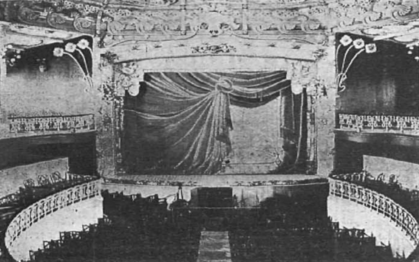 Schwarzes und weißes Foto des Auditoriums des Theatre Royal in London, das eine Bühne mit Vorhang, Geländer, ornamentierte Wände und sitzende Zuschauer zeigt, mit Text unten.
