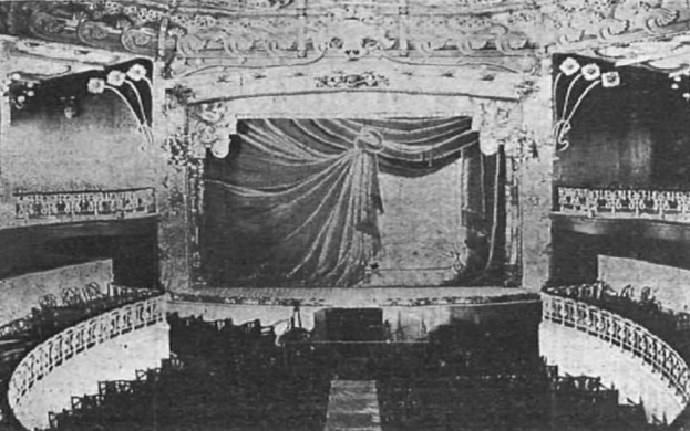 Schwarzes und weißes Foto des Auditoriums des Theatre Royal in London, das eine Bühne mit Vorhang, Geländer, ornamentierte Wände und sitzende Zuschauer zeigt, mit Text unten.