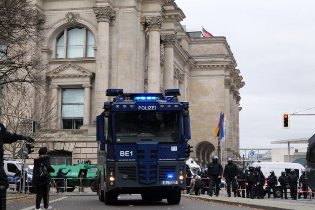 Gruppe von Polizisten vor einem großen Gebäude mit Fenstern, Säulen und Bögen, mit Fahrzeugen auf der Straße und einer Person mit einer Kamera links.