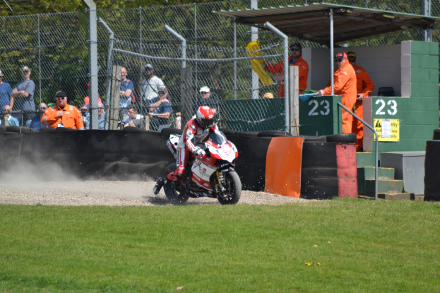 Ein Motorradfahrer in Helm und Handschuhen fährt über ein grasbewachsenes Feld, mit Zuschauern in Kappen und Schutzbrillen, einem Zaun, Treppen mit Geländern und einem nummerierten Schuppen im Hintergrund.