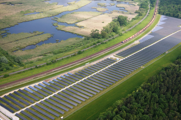 Luftaufnahme eines Solarparks mit Solarpanelen, umgeben von Bäumen, Gras, Wasser und einer nahen Bahnlinie.