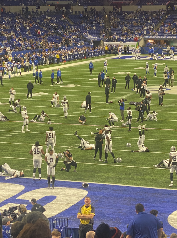 Ein Fußballspiel wird in einem Stadion gespielt, während Menschen auf dem Rasen stehen und einige auf Stühlen im Hintergrund sitzen. Eine Person hält eine Kamera, was auf eine Live-Übertragung hinweist.