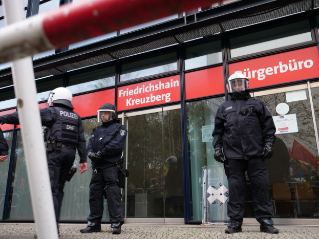 Polizisten in Uniform stehen vor einem Glasfrontgebäude, mit einem Pfahl links und Hintergrundschildern, die "Friedrichshain Kreuzberg" lesen.