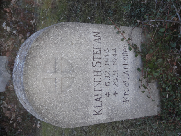 Ein Grabstein in einem grasbewachsenen Friedhof mit Pflanzen, darauf die Inschrift "Klatsch Stefan" und seine Daten.