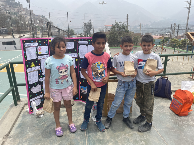 Gruppe von Kindern auf einem Basketballfeld, die Papiertüten halten, mit Vorräten auf dem Boden und einer Tafel mit Text links, vor einem Hintergrund aus Zaun, Pfosten, Straßenlaternen, Bäumen, Gebäuden, Hügeln und Himmel.