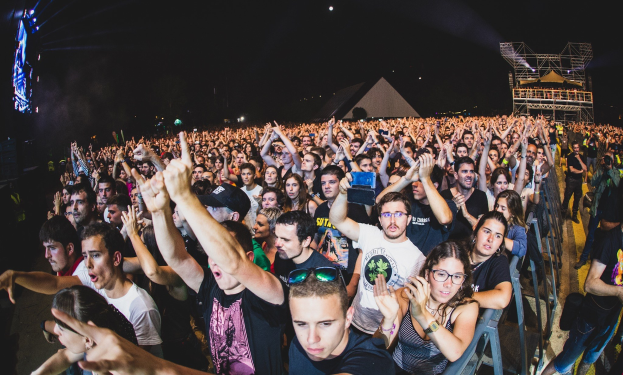 Eine große Menge Menschen auf einem Musikfestival mit erhobenen Händen, einige sitzen auf Stühlen und andere stehen, begrenzt von einem Metallzaun auf der rechten Seite, mit Gebäuden, Lichtern und einem dunklen Himmel im Hintergrund.