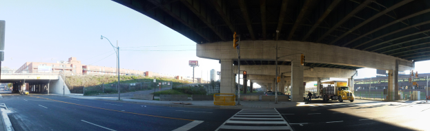 Lastwagen unter einer Brücke auf einer Stadtstraße mit Gebäuden, Straßeninfrastruktur und Fahrzeugen unter einem klaren blauen Himmel.