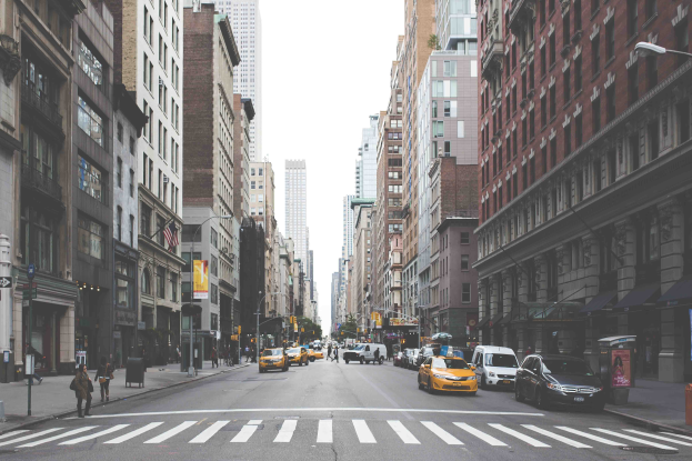 Eine belebte Stra├če in einer Stadt mit Schwerlastverkehr, Passanten, hohen Geb├Ąuden und Werbetafeln, die die Top-Attraktionen in New York City anpreisen.