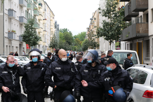 Polizisten in Masken und Helmen gehen eine Straße mit parkenden Autos entlang, flankiert von Gebäuden mit Balkonen und Geländern, mit Bäumen und einem klaren Himmel im Hintergrund.