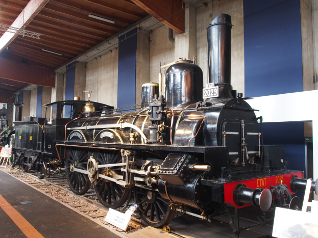 Eine große, alte Dampflokomotive in einem Museums Setting umgeben von Eisenbahnschienen, Informationsschildern und anderen Ausstellungen, mit einer Wand und Deckenbeleuchtung im Hintergrund.