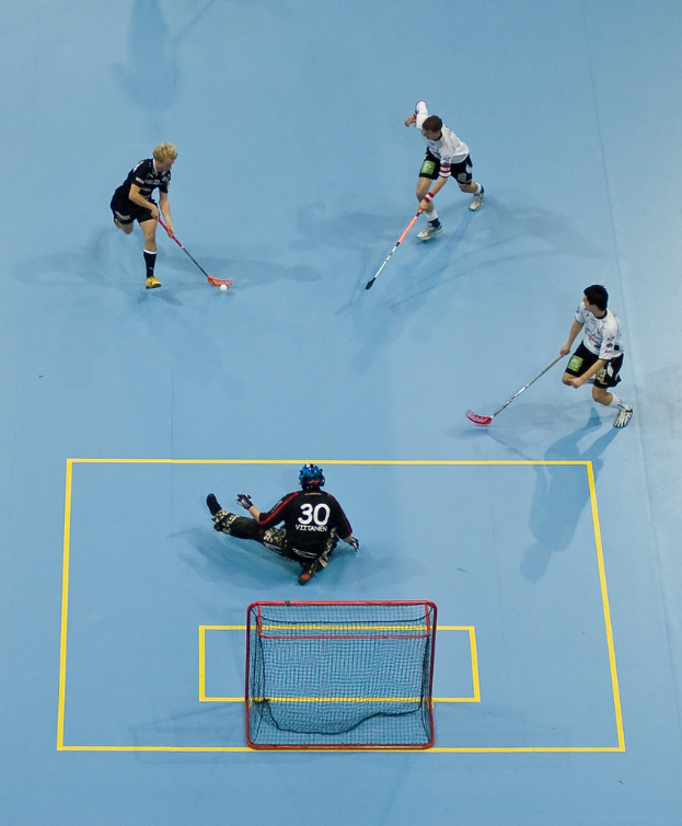 Drei Personen in Sportbekleidung spielen Hockey, wobei eine Person rechts einen Ball mit einem Hockey-Schläger schlägt und eine Person hinter einem Netz auf dem Boden sitzt.