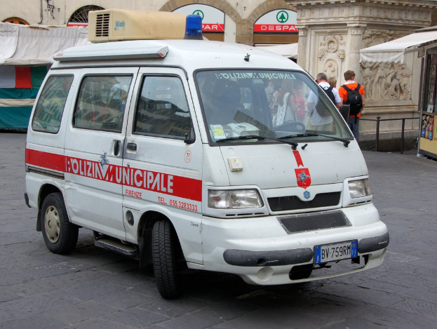 Ein weißer Van fährt auf einer Straße, während eine Gruppe von Menschen daneben steht, ein Schuppen auf der rechten Seite und Gebäude und Metallstangen im Hintergrund.
