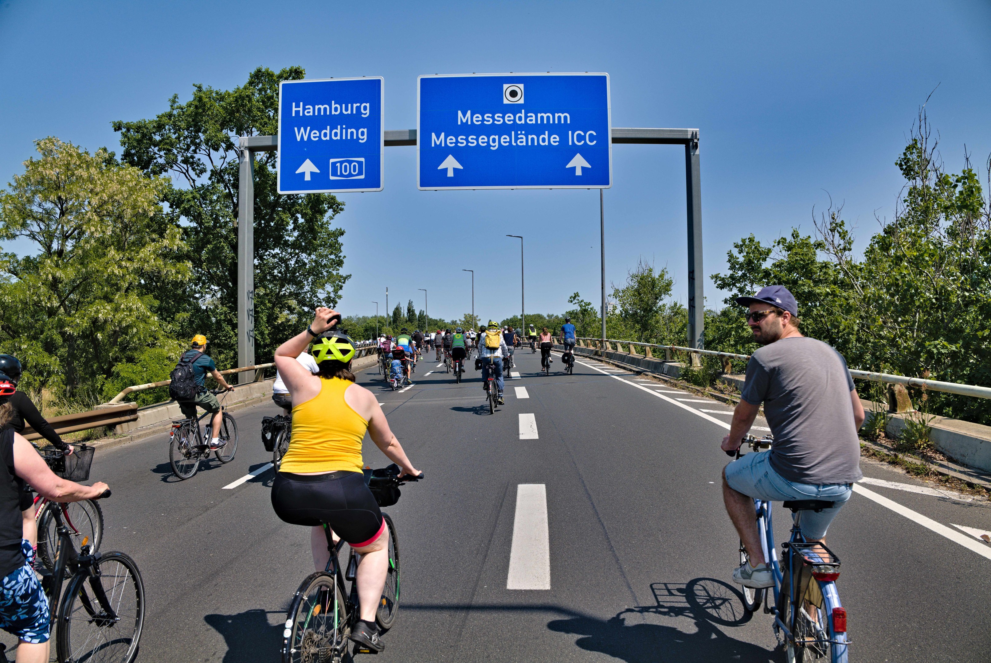 Gruppe von Radfahrern mit Helmen auf einer Straße mit einer Begrenzung auf einer Seite und Bäumen auf der anderen, Laternen im Hintergrund unter einem klaren blauen Himmel, mit einem Schild, das eine Radtour in Hamburg oben anzeigt.