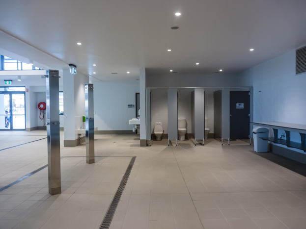 Große Öffentliche Toilette mit mehreren Toiletten, tragenden Säulen, einem Mülleimer, einer Tür, einer Schilderwand, einem angebrachten Feuerlöscher und Deckenleuchten.