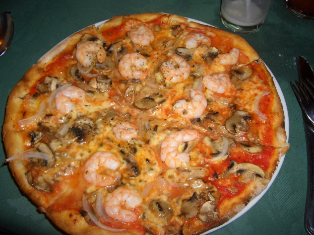 Eine Pizza mit Pilzen, Garnelen und anderen Belägen auf einem Teller, begleitet von einer Gabel, einem Messer und einem Löffel, mit zwei Gläsern davor.