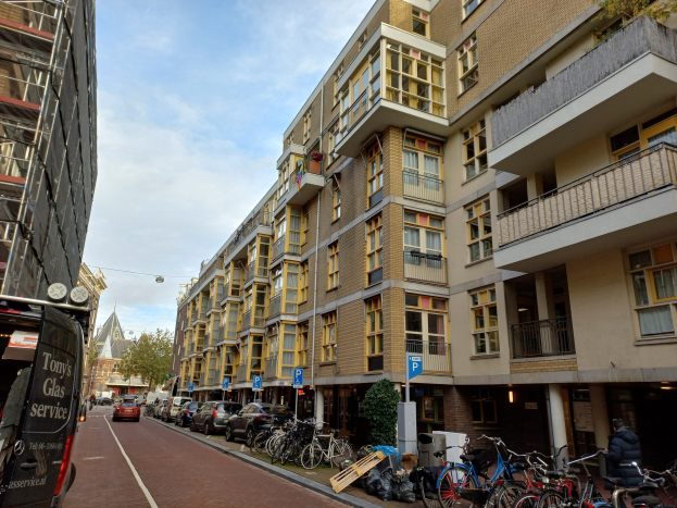 Eine Straße in Amsterdam mit parkenden Fahrrädern, fahrenden Fahrzeugen, Gebäuden, Schildern, Bäumen und einem bewölkten Himmel.