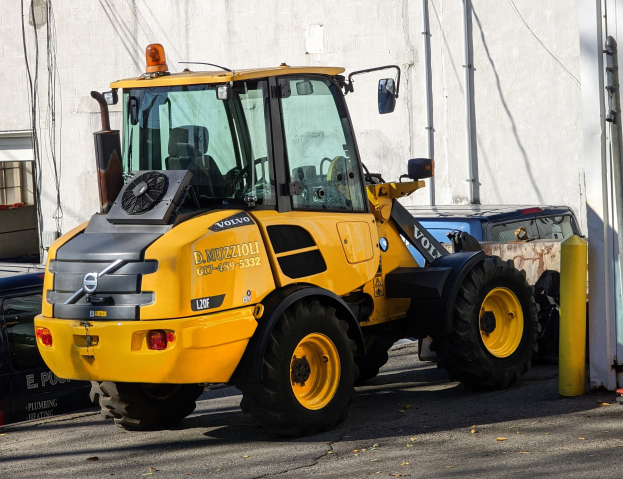 Ein gelber Radlader auf einer Straße vor einem Gebäude mit einer Wand im Hintergrund geparkt, mit anderen Fahrzeugen in der Nähe.
