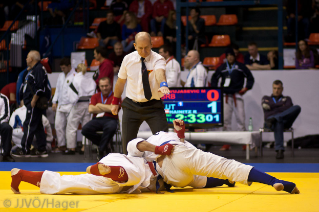Zwei Judoka kämpfen in der Mitte einer Begegnung, während ein Schiedsrichter daneben steht und Zuschauer von Stühlen und Stehplätzen sowie Geländern, Treppen und einem Bildschirm mit Zahlen und Text im Hintergrund zusehen.