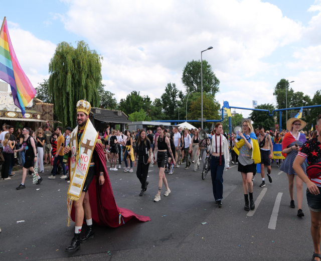 Eine Gruppe von Menschen marschiert bei der Pride Parade 2018, einige mit Musikinstrumenten und Mützen, die eine Regenbogenfahne tragen, auf einer Straße mit Laternenmasten, Bäumen und Hütten unter einem bewölkten Himmel.