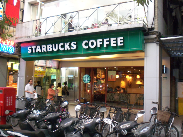 Ein Starbucks Kaffeehaus in Hong Kong mit Motorrädern vorne, einem roten Briefkasten links, Menschen rechts und einem Fahrrad und Rad in der Mitte.
