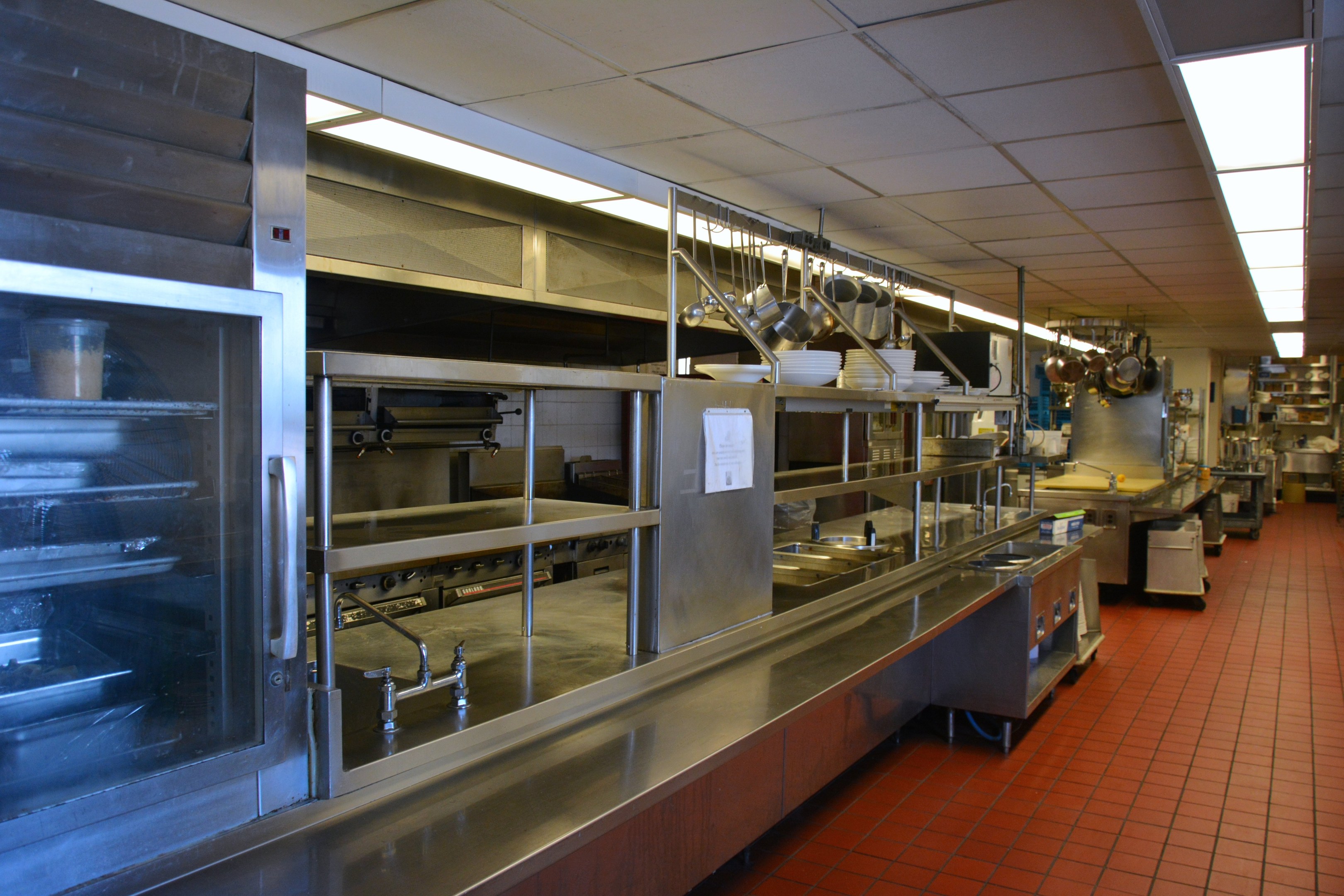 Gewerbeküche mit Edelstahl-Theke, Deckenleuchten, hängenden Utensilien, einem Spülbecken, Wasserhähnen, Schränken und Gegenständen auf Regalen.