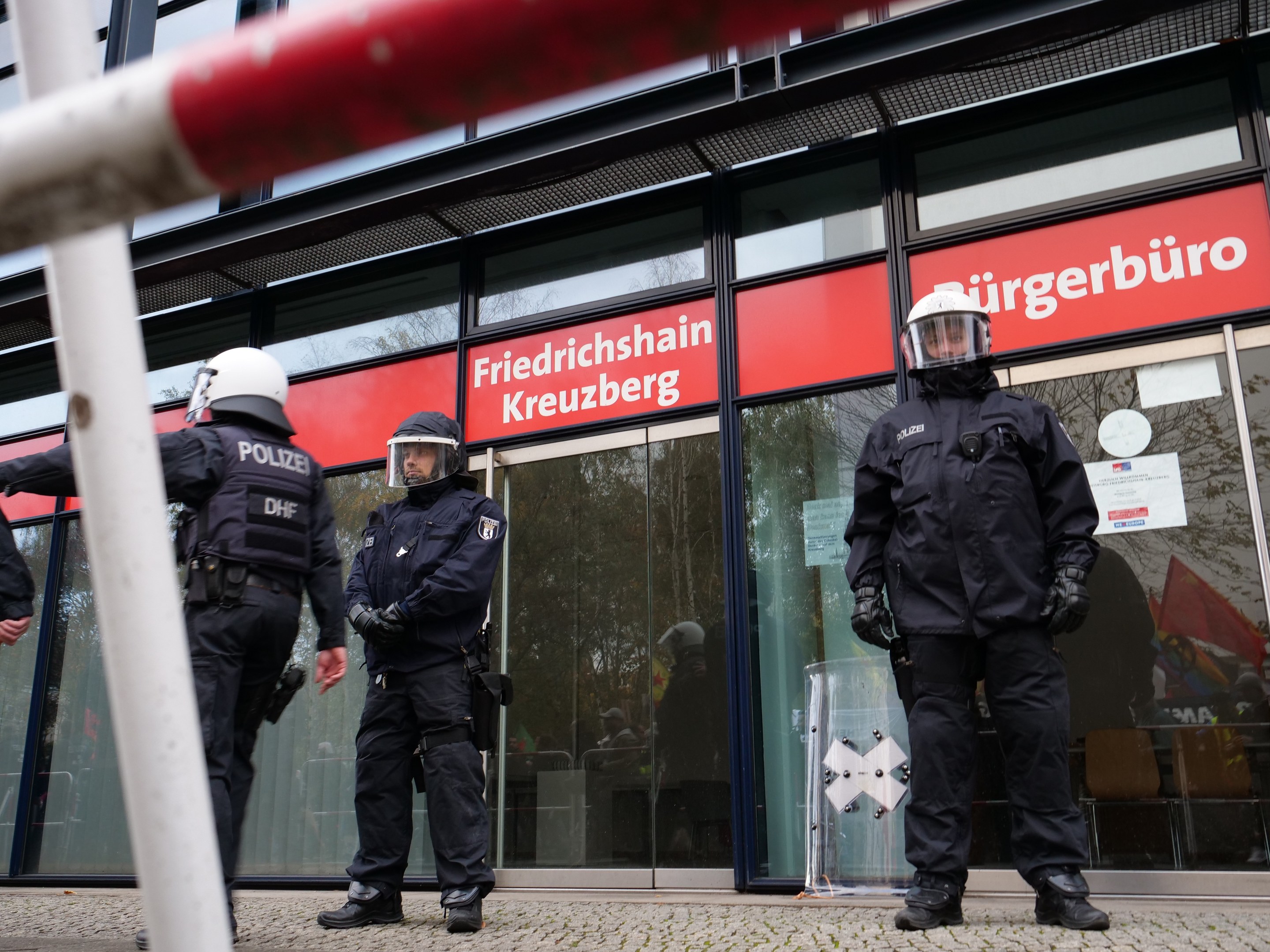 Polizeibeamte in Uniform stehen vor einem Glasgebäude, tragen Helme und Handschuhe, mit einer Stange links und Schildern im Hintergrund, die "Friedrichshain Kreuzberg" lesen.