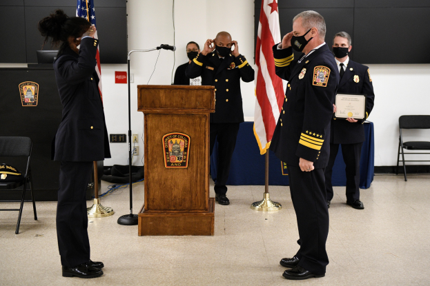 Gruppe uniformierter Personen in Masken vor einem Rednerpult bei einer Amtseinführung für einen neuen Feuerwehrchef, der eine Urkunde hält, mit Flaggen, Stühlen, Tischen und einer Wand im Hintergrund.